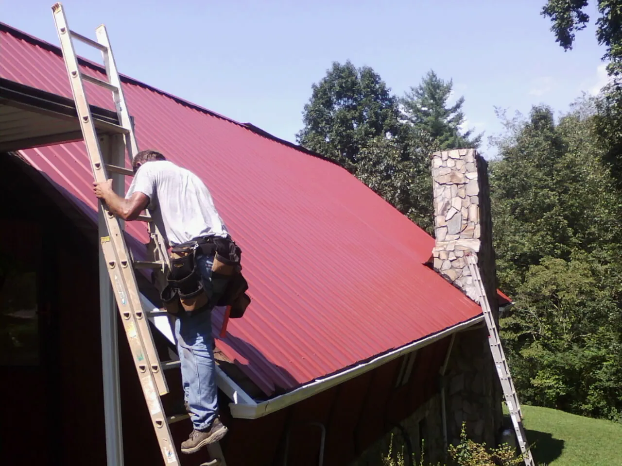 Roofer on a ladder beside a red metal roof and stone chimney — Soffit & Fascia Repair by Roofing Brothers in Morgan City